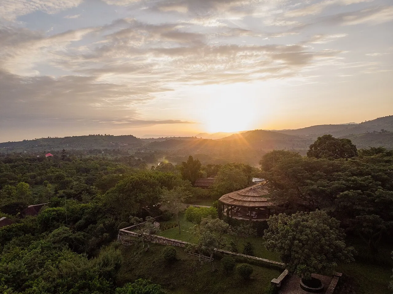 Aerial view of Kyambura Gorge Lodge's round thatched building among trees with the sun low over hills