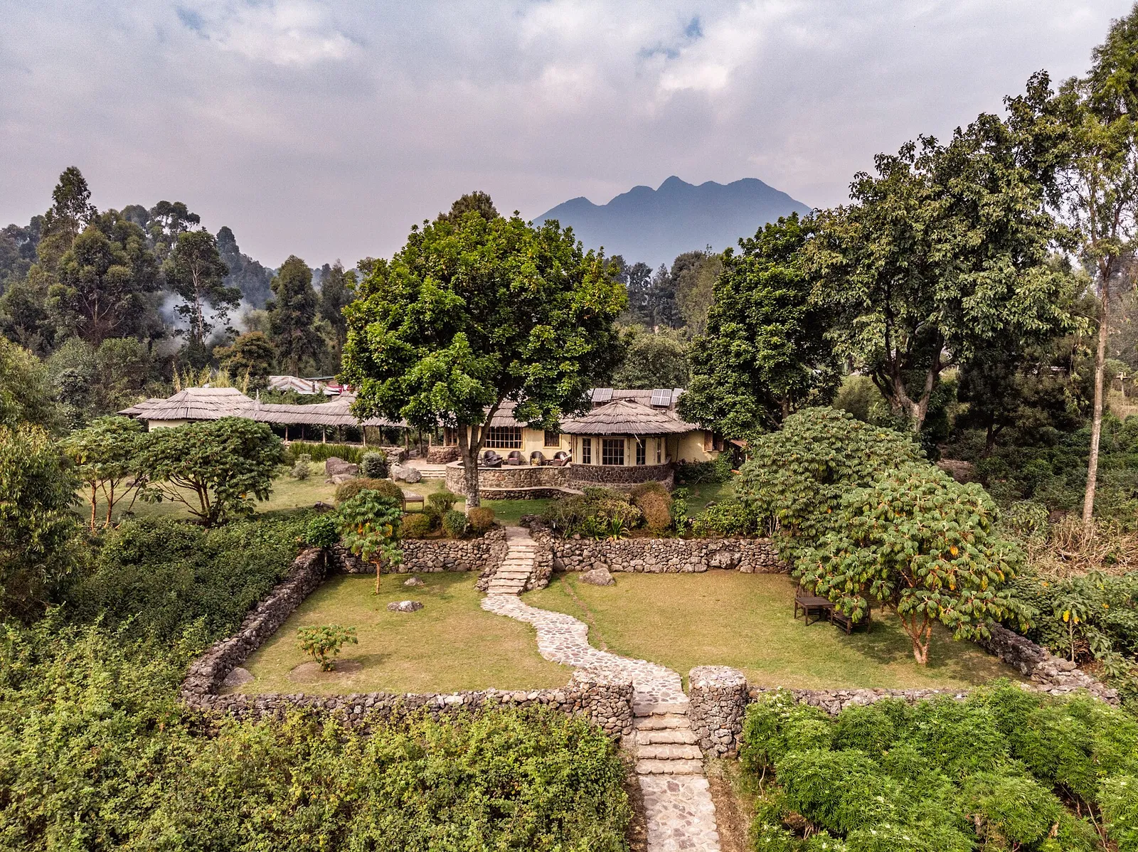 Mount Gahinga Lodge's thatched main building and stone-walled garden among trees, with a volcanic peak behind, Mgahinga, Uganda