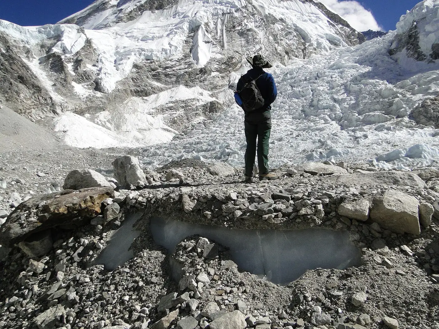 A trekker in a wide-brimmed hat standing on the rocky Khumbu Glacier at Everest Base Camp, the icefall behind him