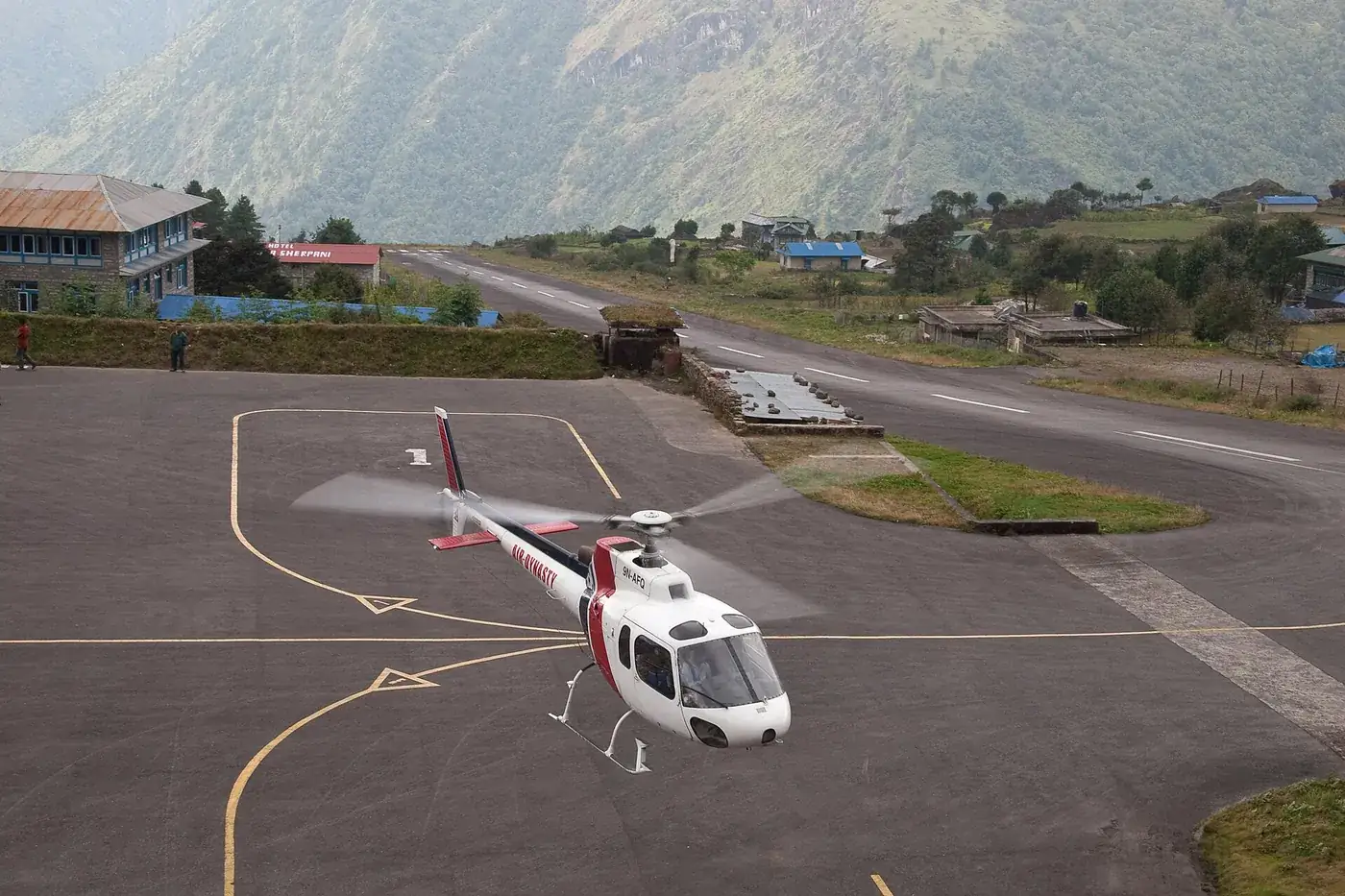 A white and red helicopter landing on the apron at Lukla airport, with the sloping runway and green hills behind
