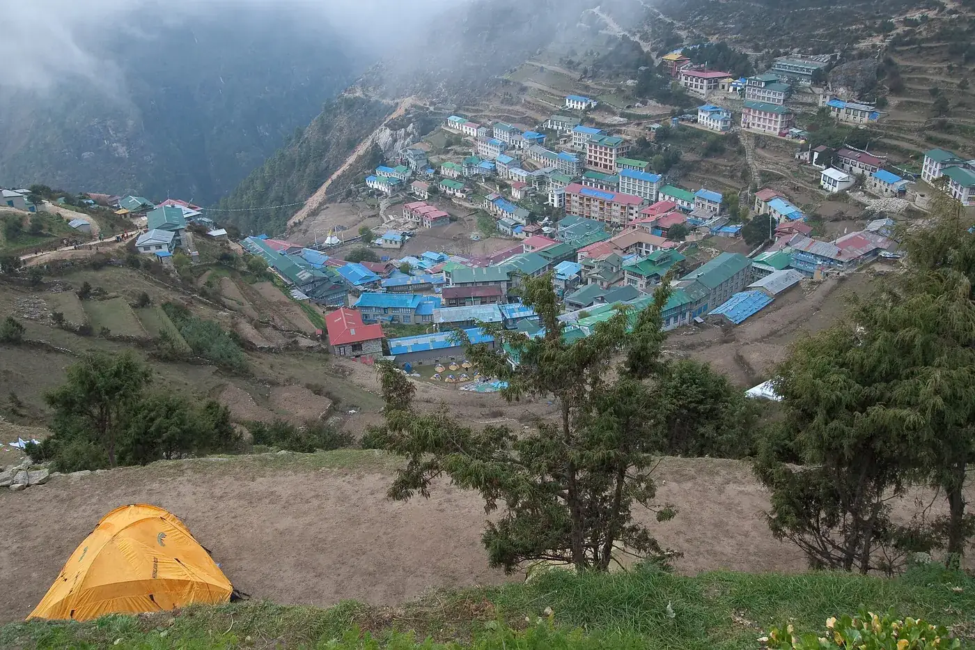 Namche Bazaar’s blue and green roofs spread across a hillside bowl, seen from above with an orange tent in the foreground