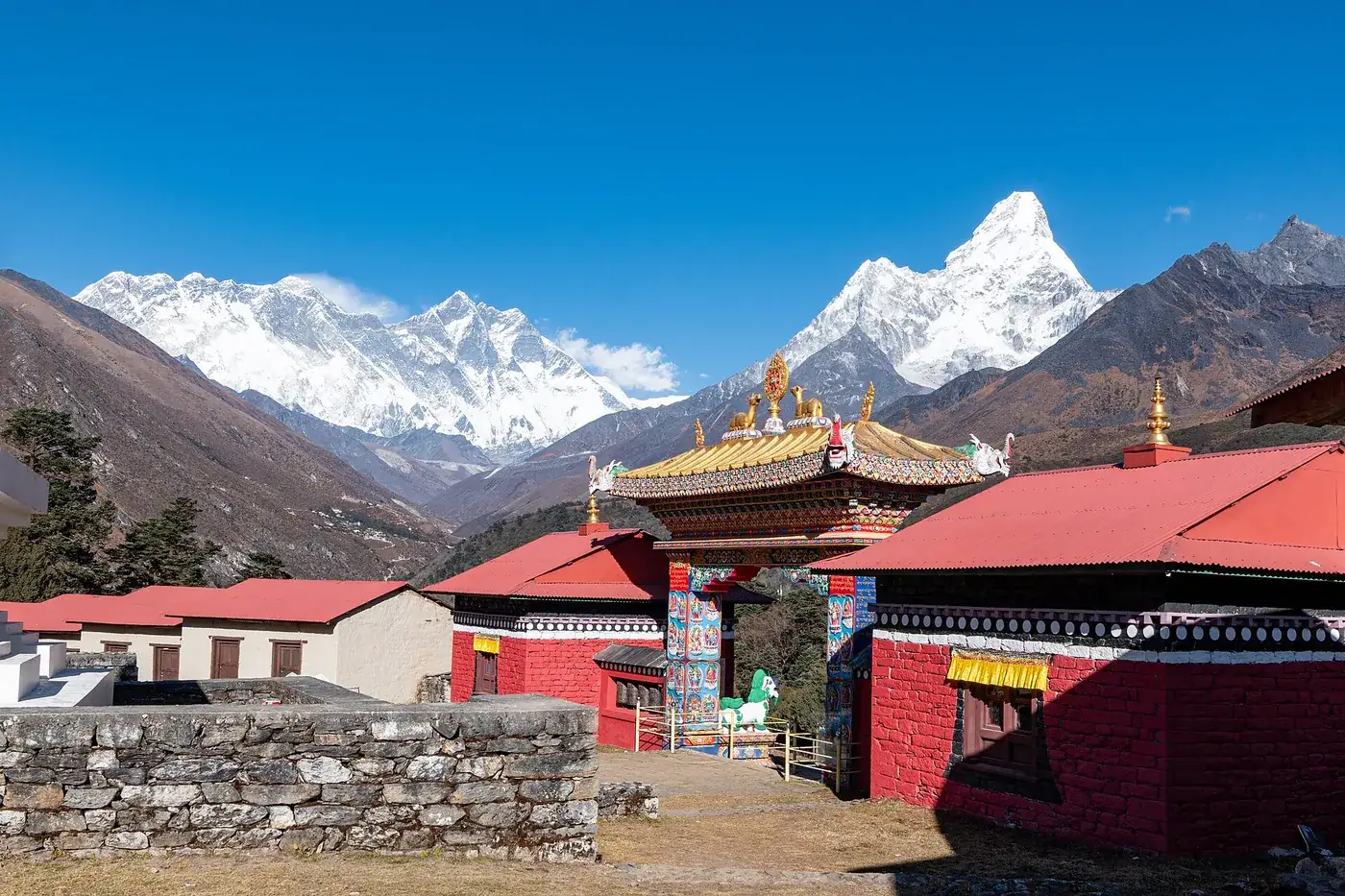 The painted entrance gate and red buildings of Tengboche Monastery under a blue sky, with snow peaks behind