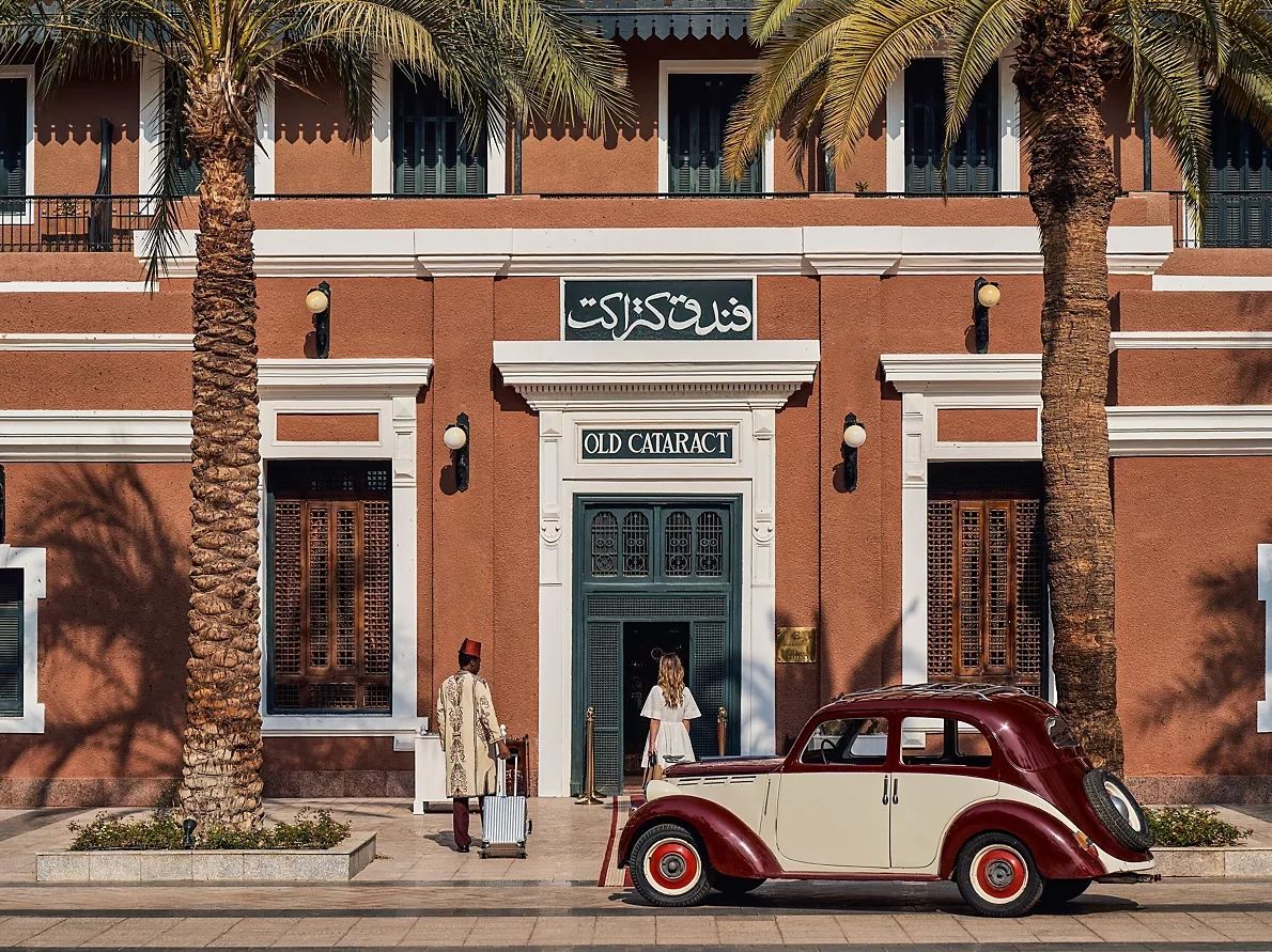 A doorman in a red tarboosh beside a suitcase at the terracotta entrance of the Old Cataract hotel in Aswan, with a vintage maroon car in front
