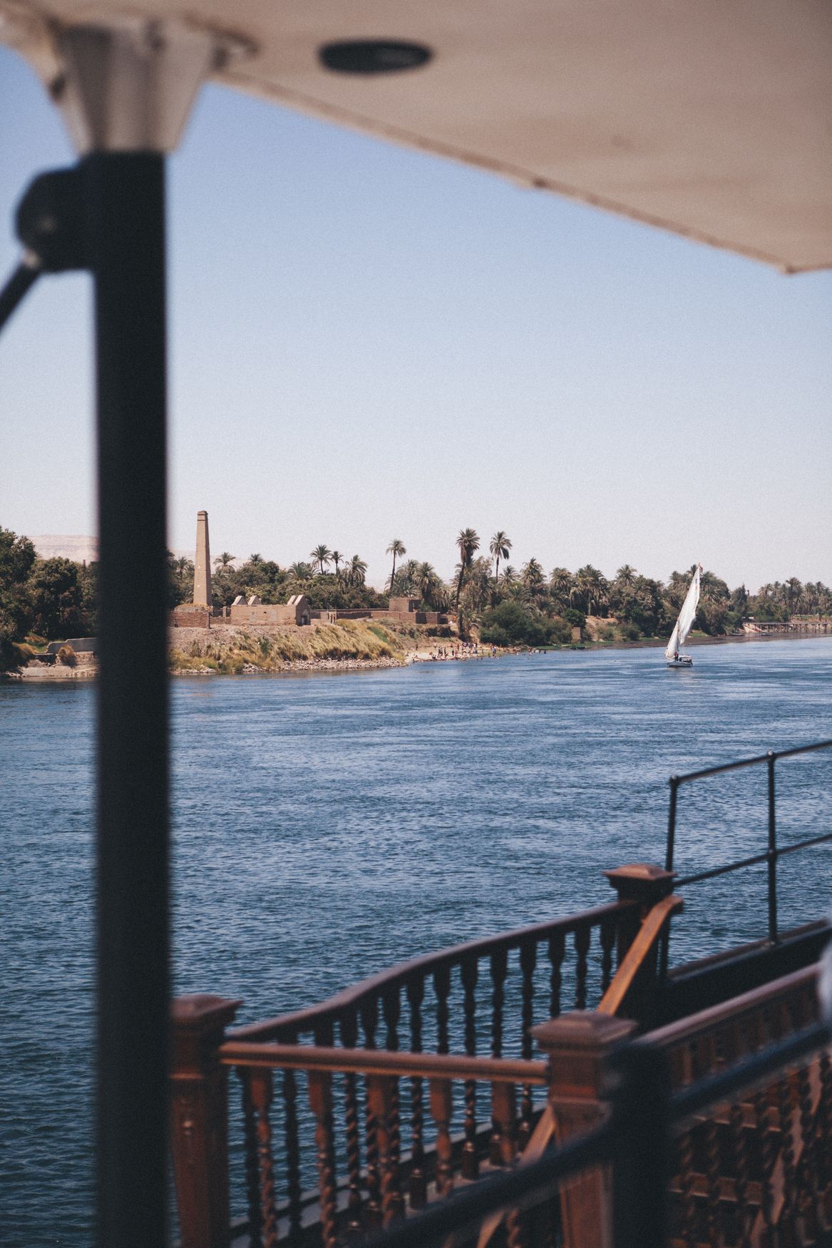 The view from the shaded deck of the Steam Ship Sudan, with its wooden rail, across the Nile to a white-sailed felucca and palms on the far bank