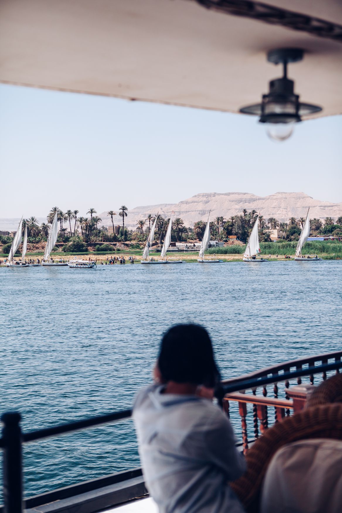 A traveller seen from behind at the rail of the Sudan’s shaded deck, looking across the Nile at a line of feluccas and desert hills