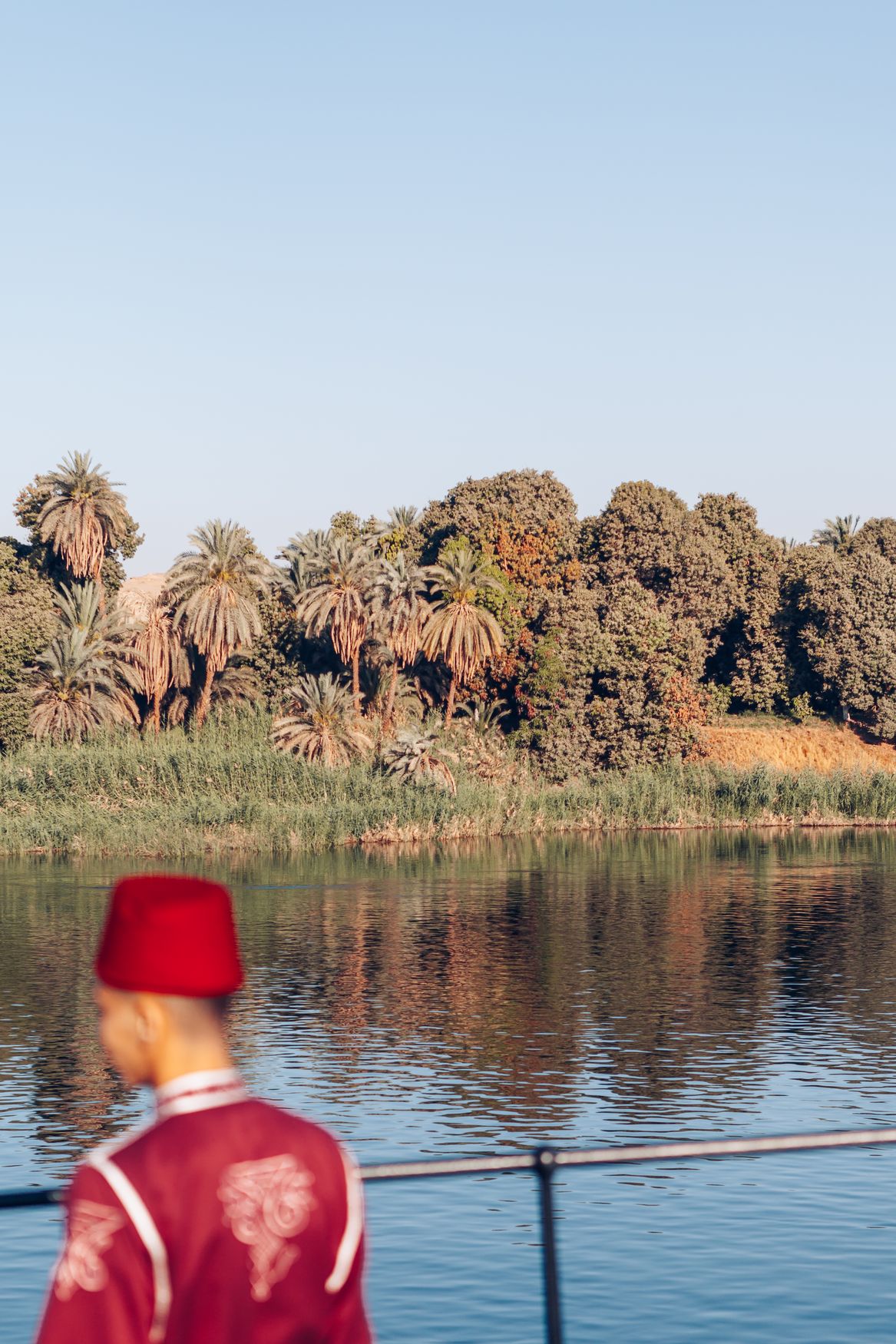 A crew member in a red tarboosh and embroidered jacket at the rail of the Steam Ship Sudan, with palms on the riverbank behind