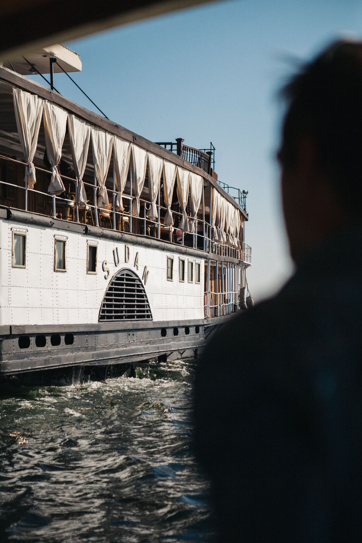 The white paddle steamer Sudan seen from the deck of another boat, its name painted on the paddle housing, with a passenger in silhouette