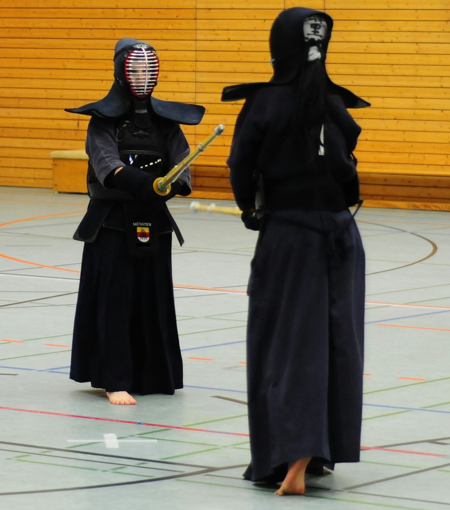 Two people in kendo armour and helmets holding bamboo swords in a sports hall