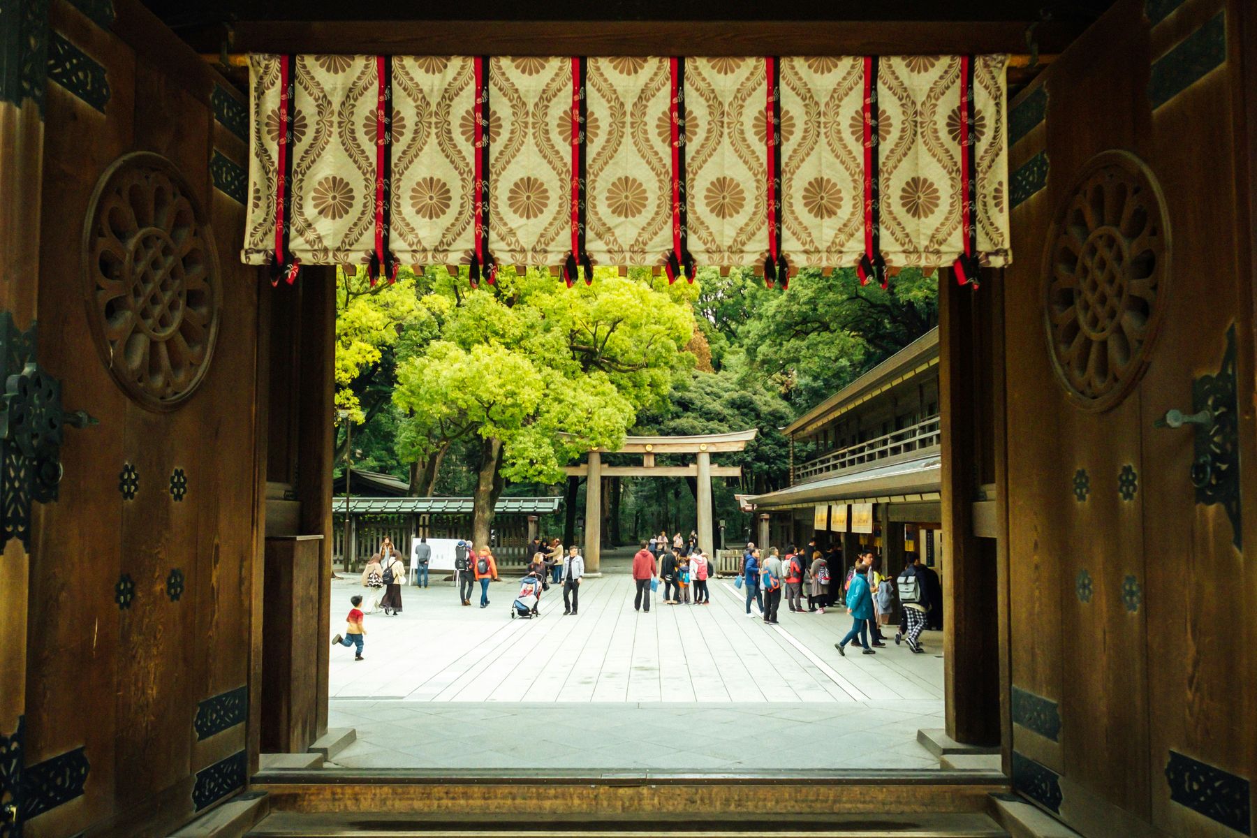 A shrine courtyard seen through a carved wooden gate hung with a patterned curtain, with a torii gate and visitors beyond