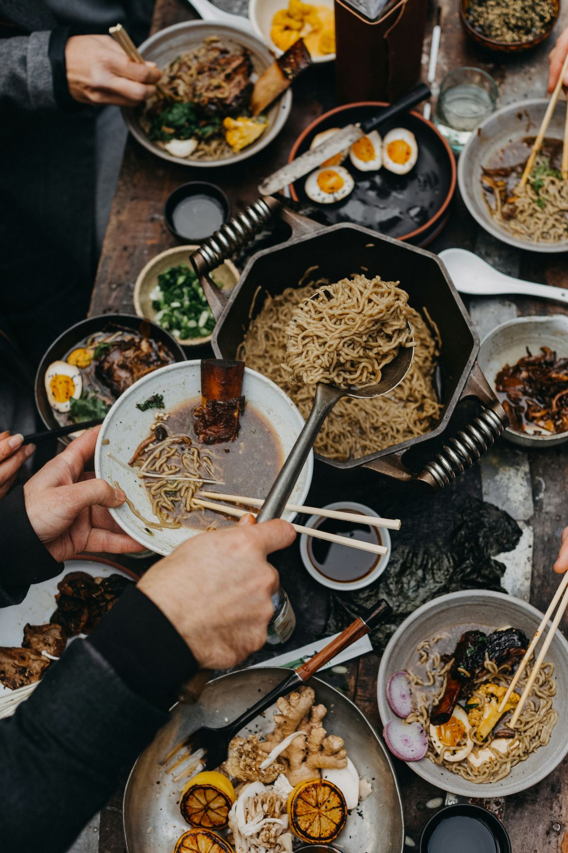 Bowls of ramen with eggs, chopsticks and side dishes spread across a wooden table, seen from above