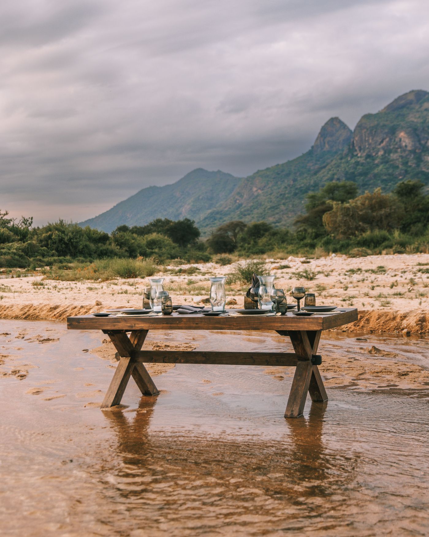 A dining table laid with crockery set in a shallow river with mountains behind at Kalepo Camp, Kenya