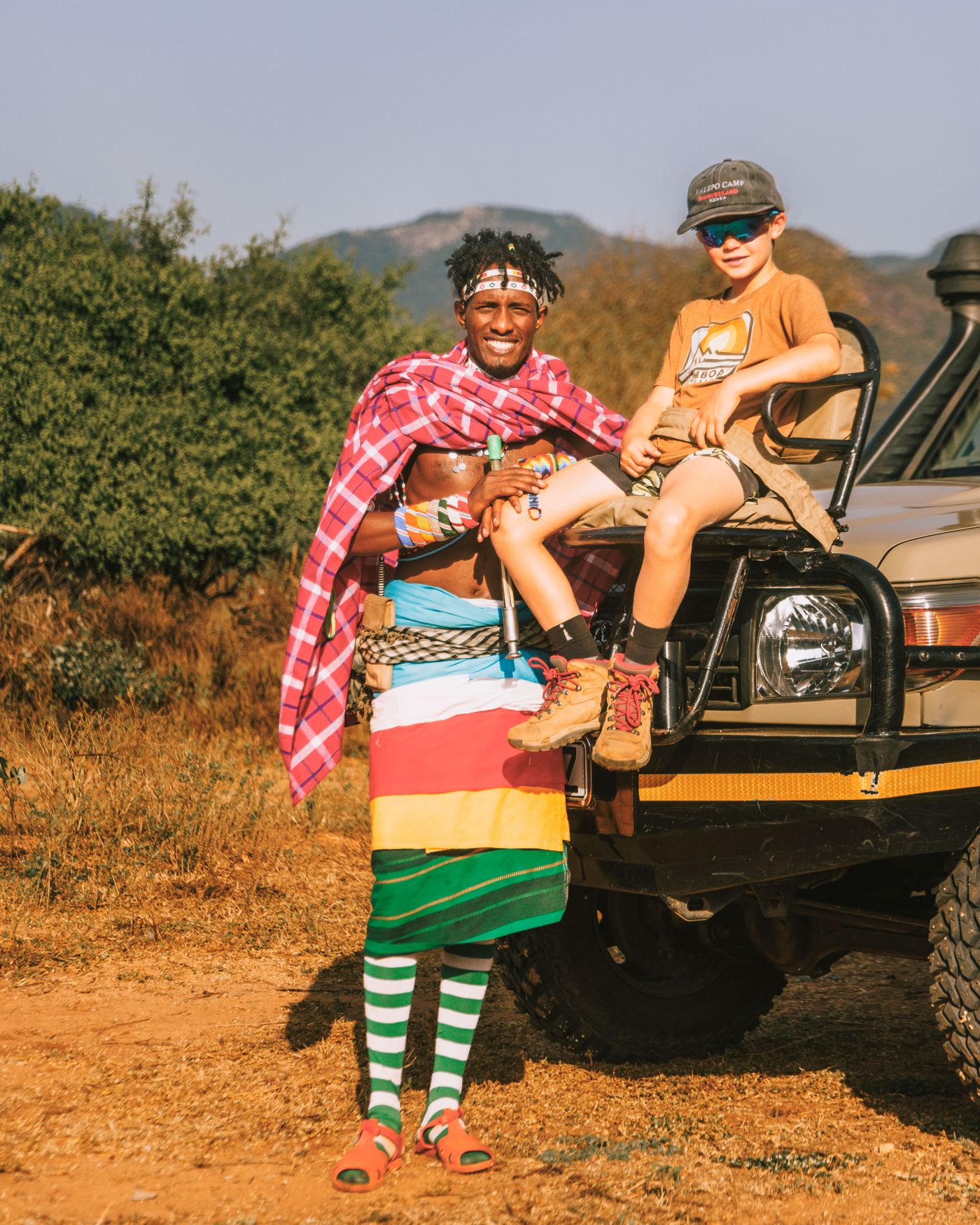 A Samburu guide in red and green robes holds a smiling child wearing a hat on the bonnet of a safari vehicle at Kalepo Camp, Kenya