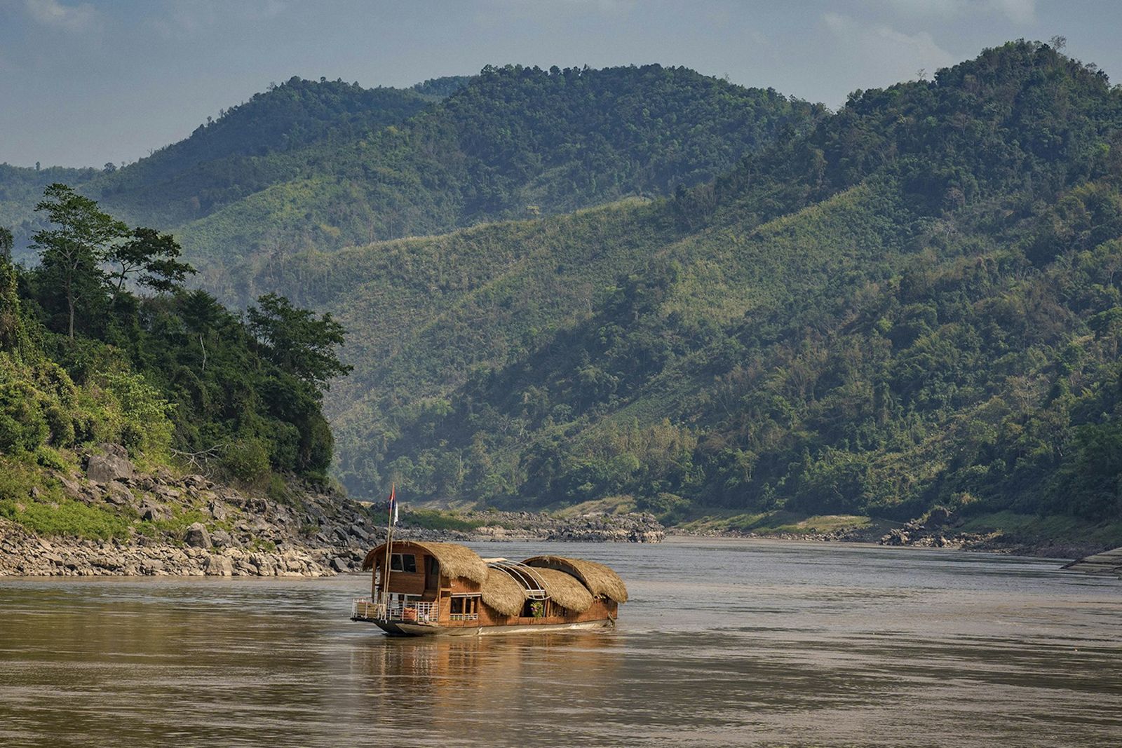 A wooden riverboat with a thatched roof on the brown water of the Mekong, with forested hills behind