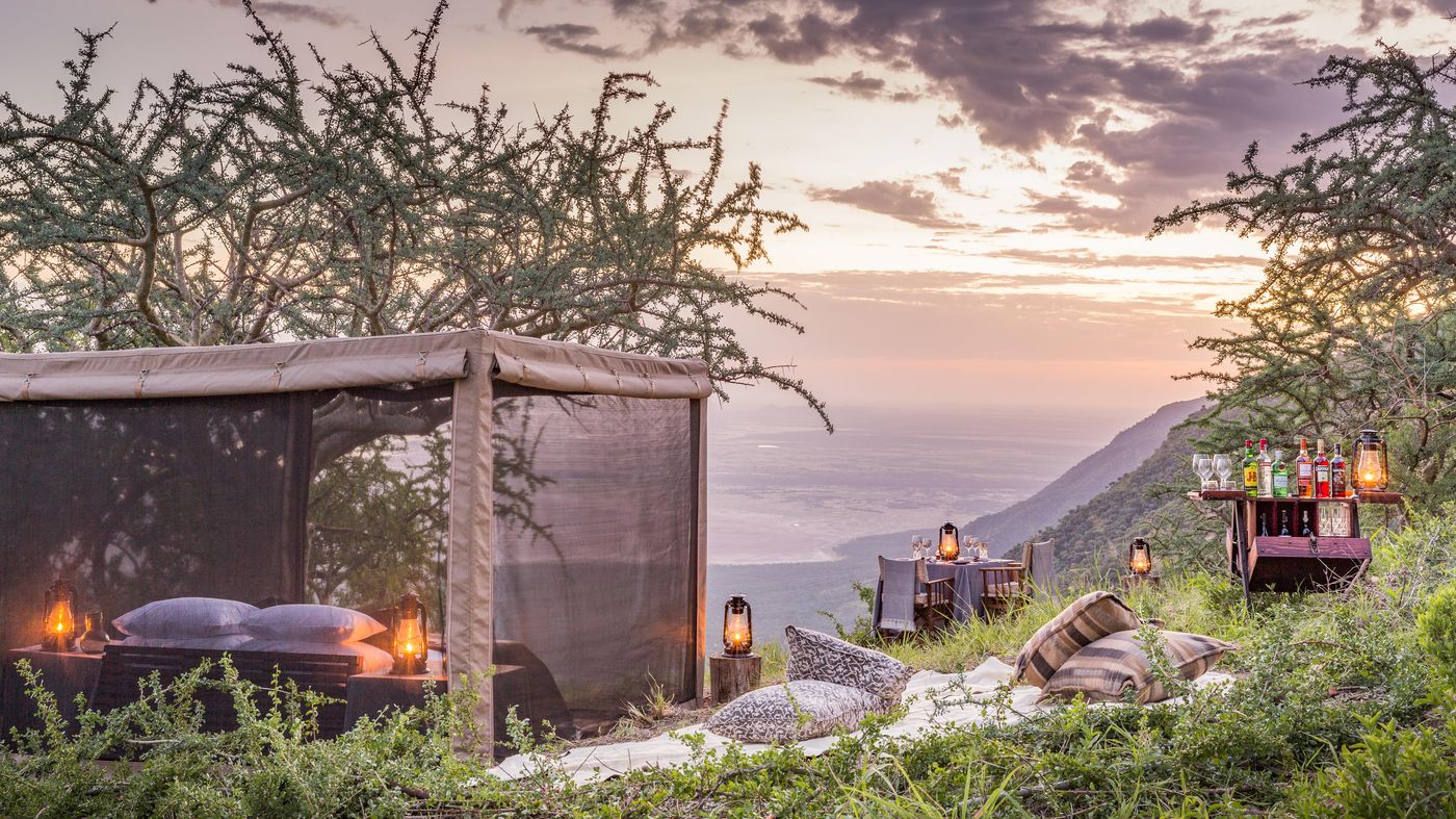 A fly-camp bed under a mesh canopy on an escarpment at sunset with lanterns and a dining table, Mwiba Lodge, Tanzania