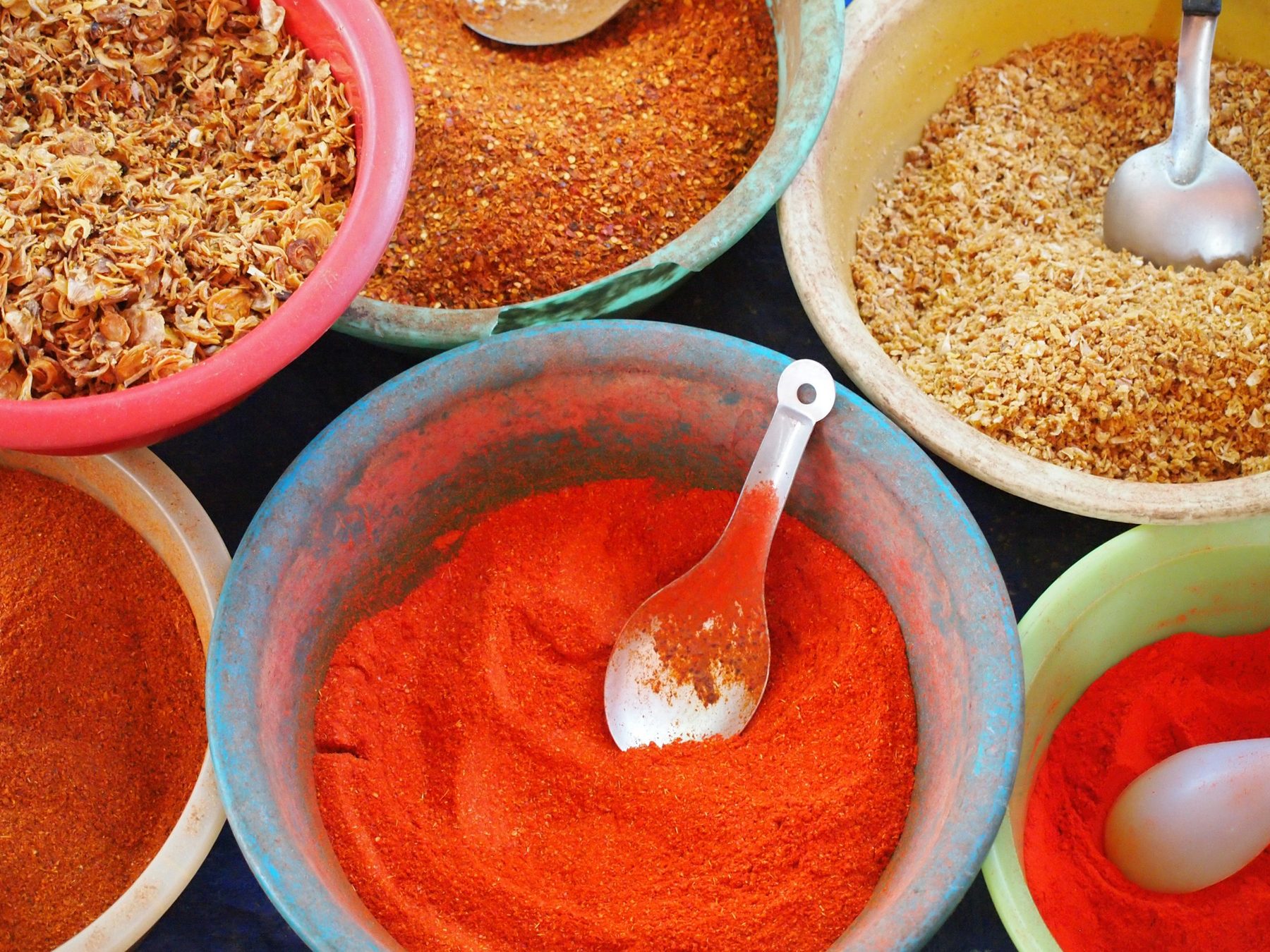 Plastic bowls of ground chilli, dried flowers and spices with metal scoops, seen from above at a market stall