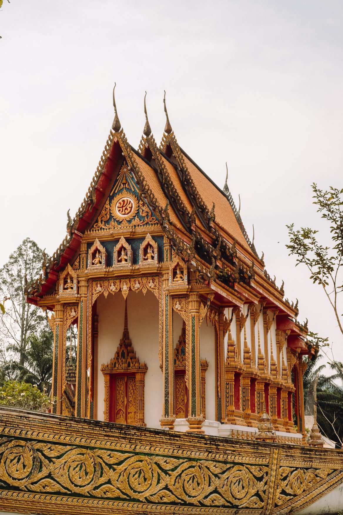 A Lao temple with a steep red tiled roof, gilded gables and gold-patterned columns, with a carved balustrade in the foreground