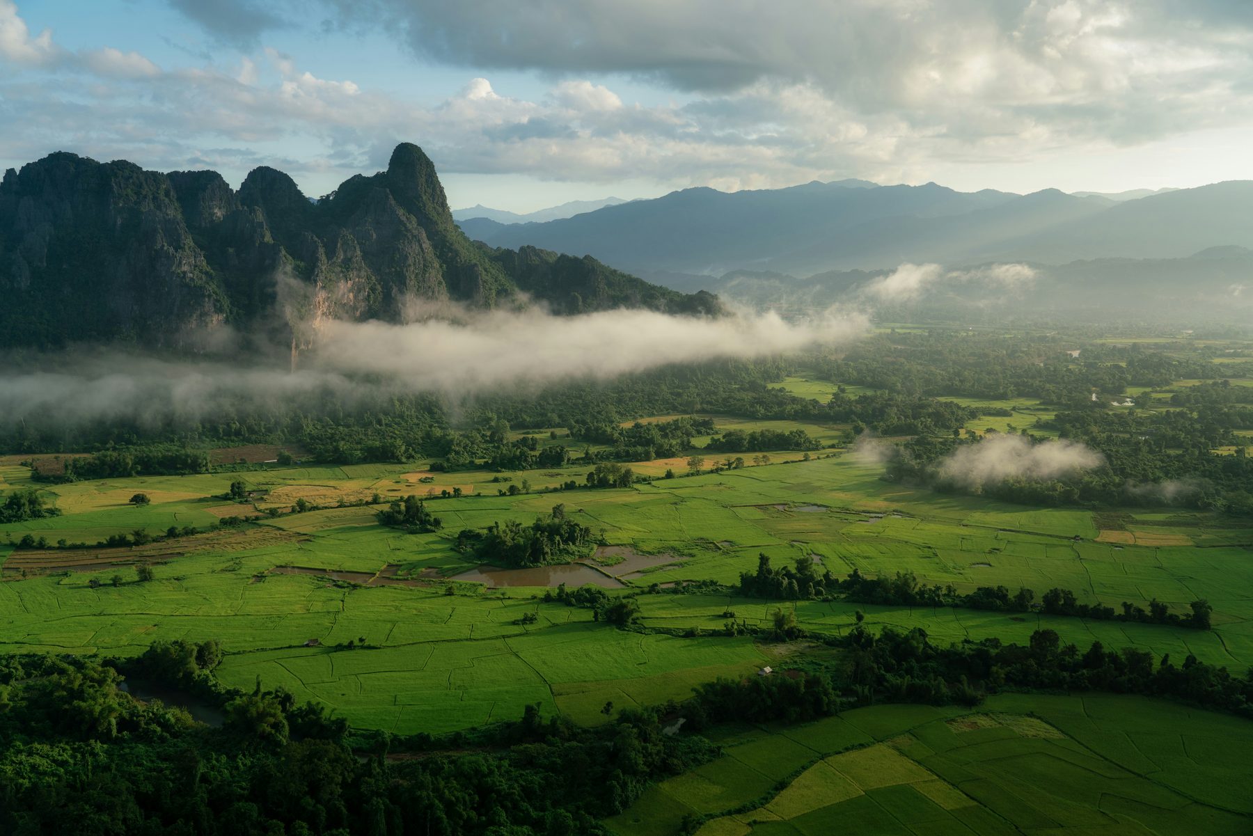 Limestone mountains rising above green rice fields under low cloud and mist near Vang Vieng, Laos