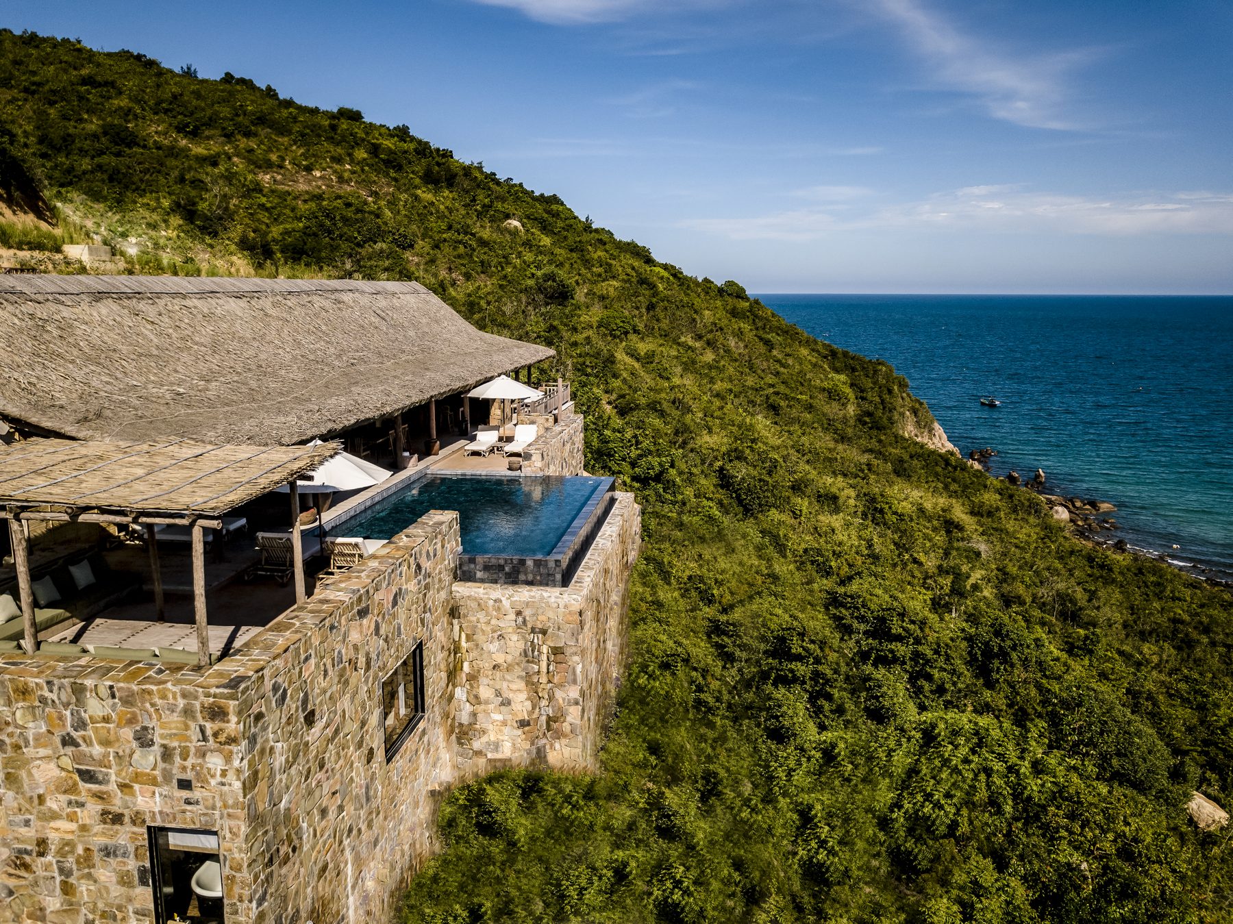 A stone villa with a thatched roof and an infinity pool on a green hillside above a dark blue sea