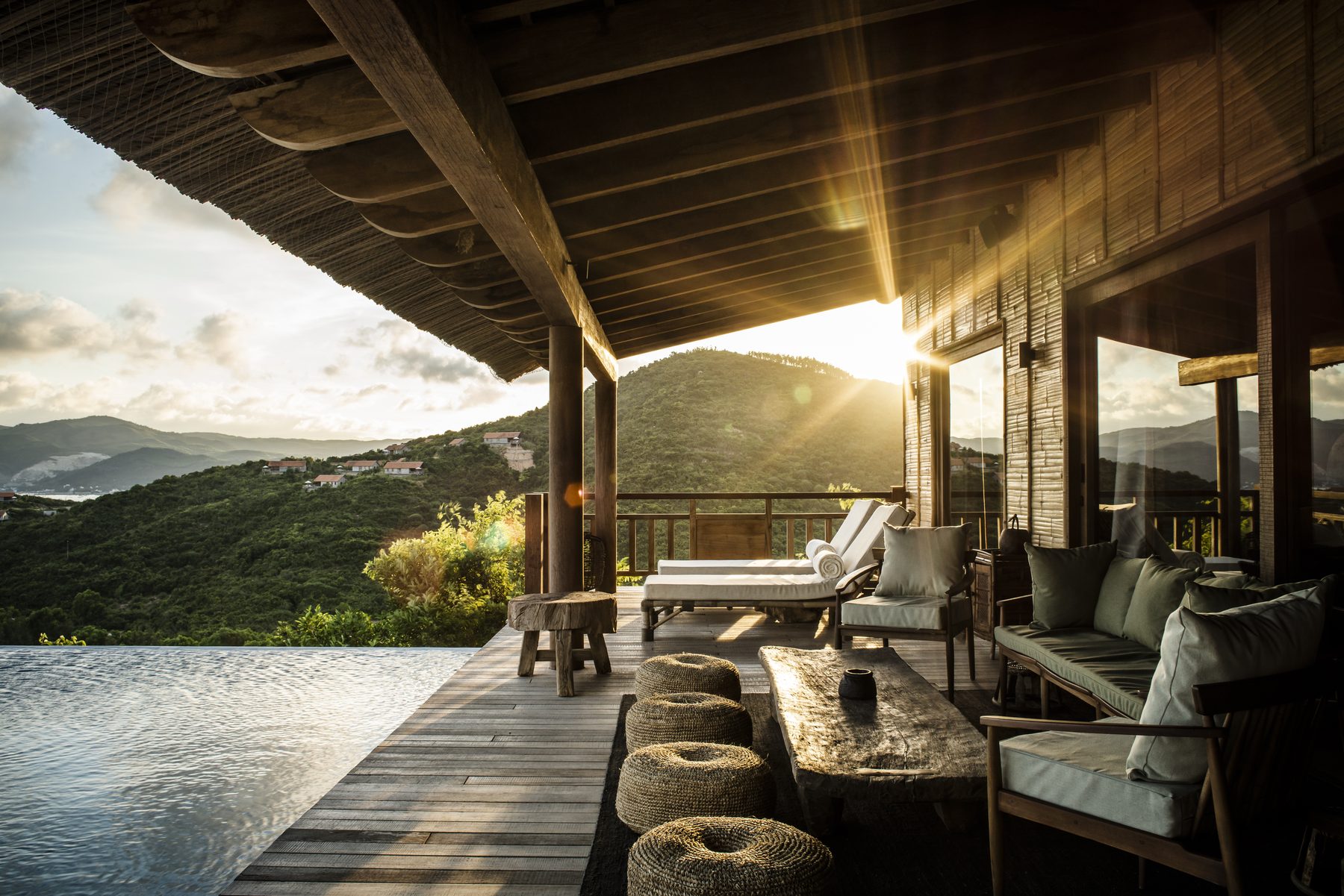 A covered villa terrace with loungers and sofas beside a pool, looking out over a bay at sunset