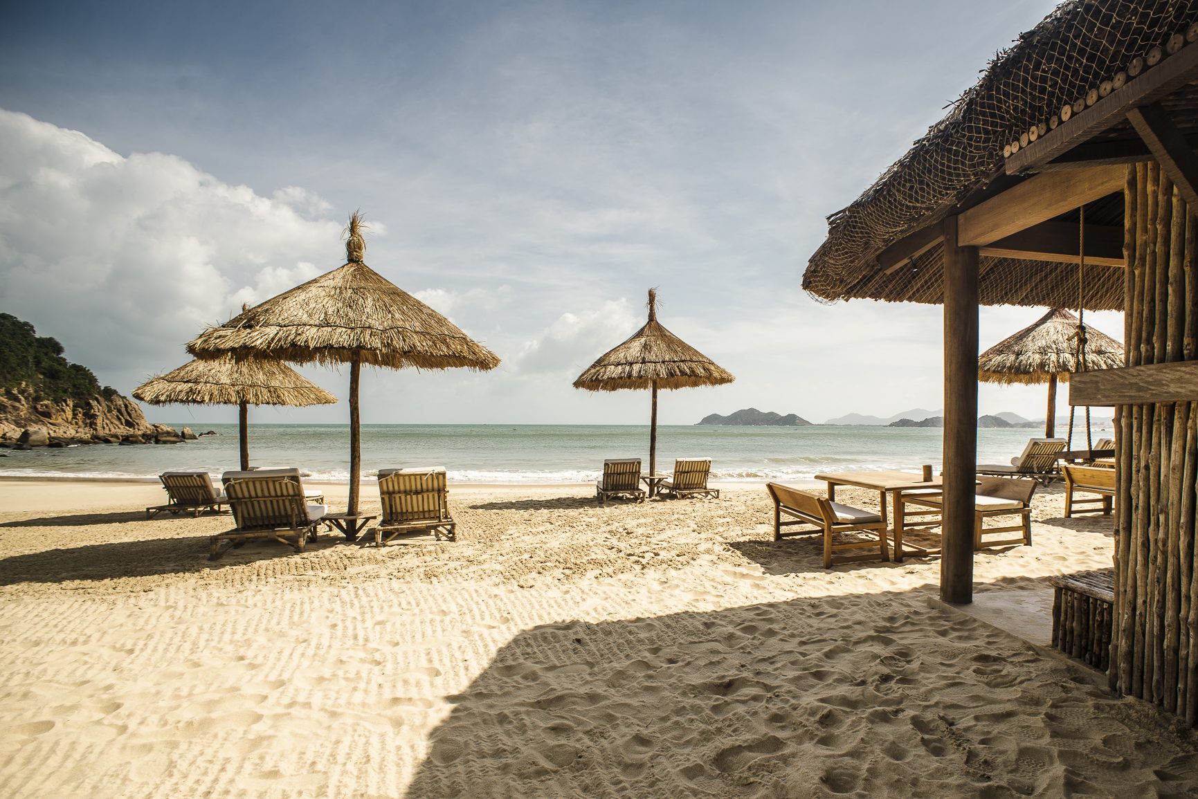 Thatched beach umbrellas and wooden loungers on pale sand under a cloudy sky, with hills across the water
