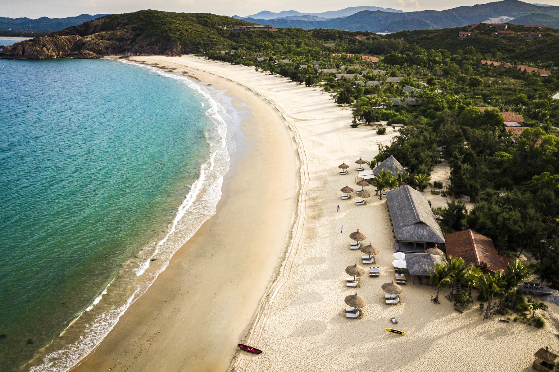 A curved sandy bay with turquoise water, thatched umbrellas on the beach and green hills behind, seen from above