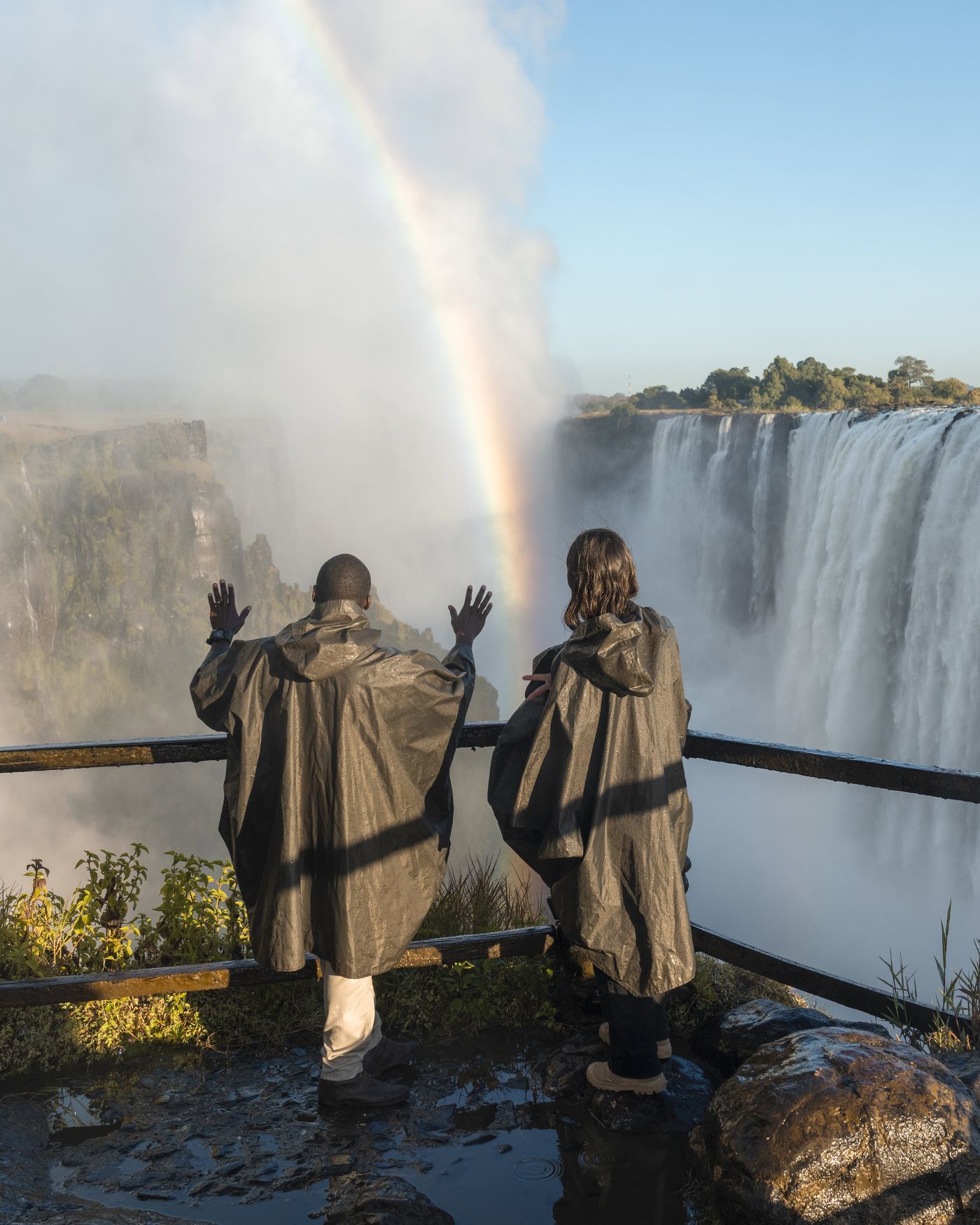 Two people in rain ponchos stand at the edge of Livingstone Island with the Victoria Falls and a rainbow in the spray behind them