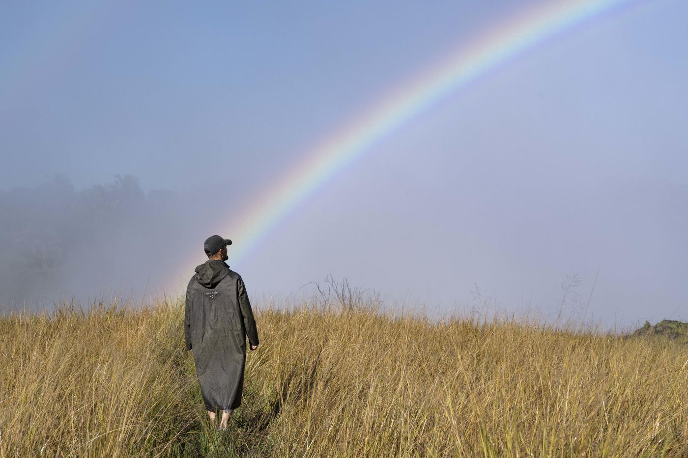 A guide walks through long grass under a full rainbow in the spray of Victoria Falls, Zambia