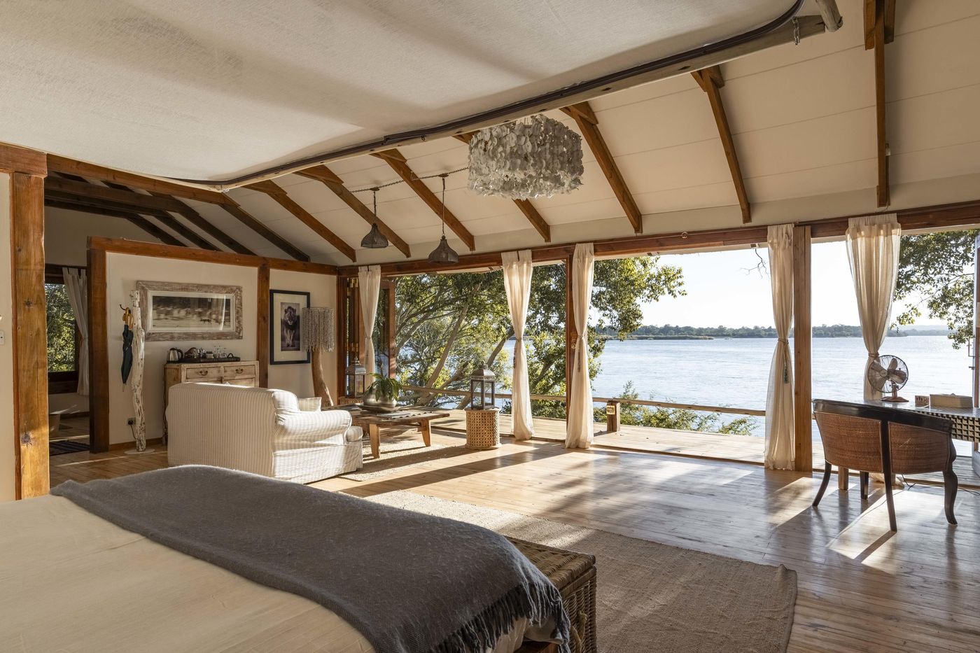A bedroom at Tongabezi Lodge open on one side to the Zambezi River, with a wooden desk, white bed and a river view
