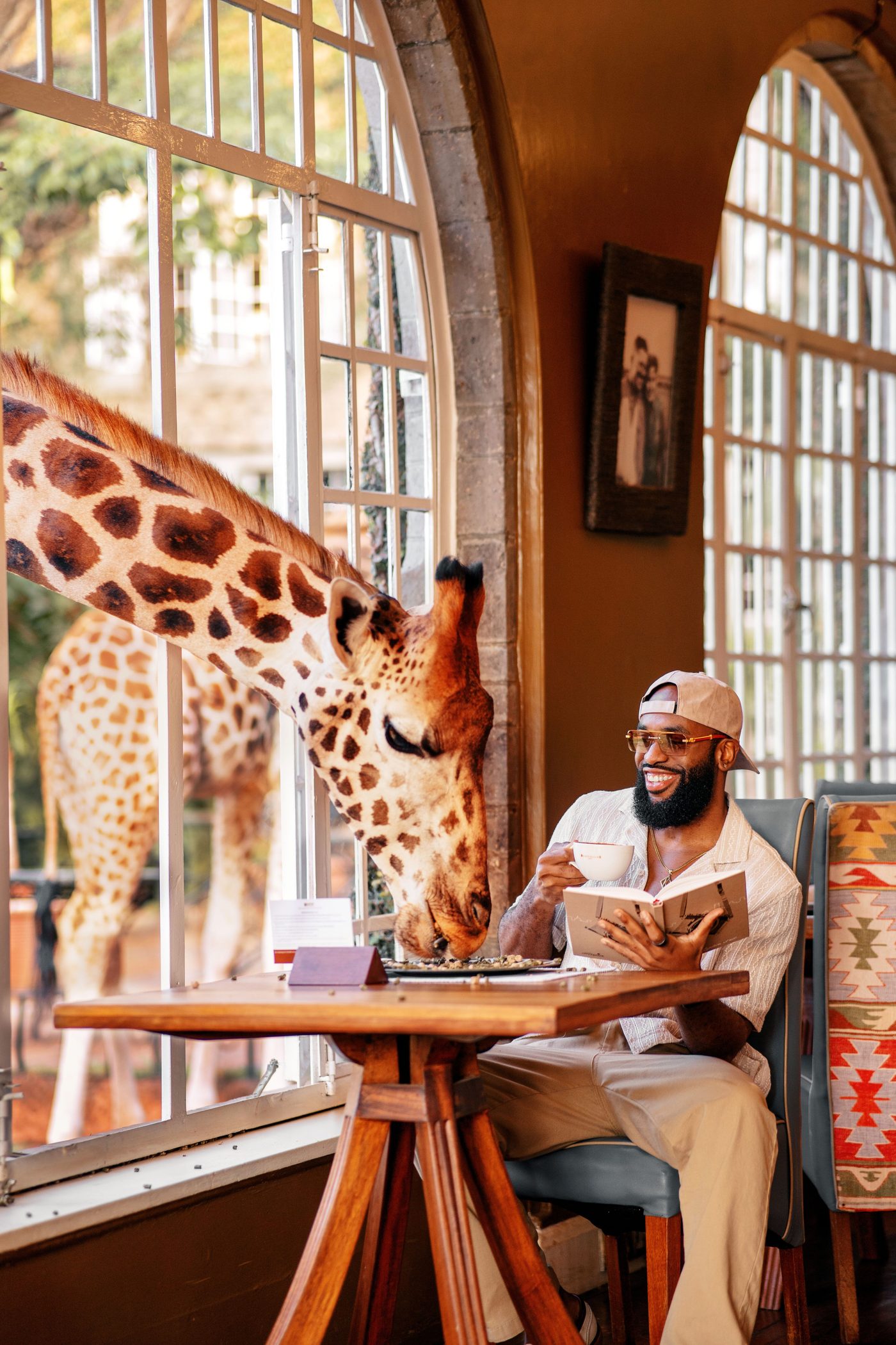 A man in a cap reads at a breakfast table while a giraffe leans in through the arched window at Giraffe Manor, Nairobi