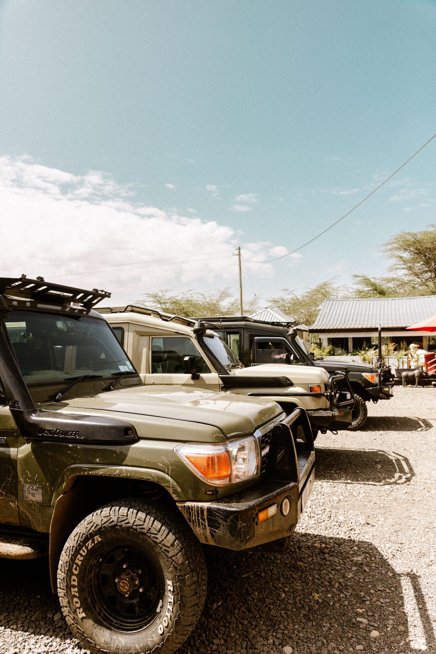 A row of green Toyota Land Cruiser safari vehicles parked on gravel outside a lodge under a blue sky