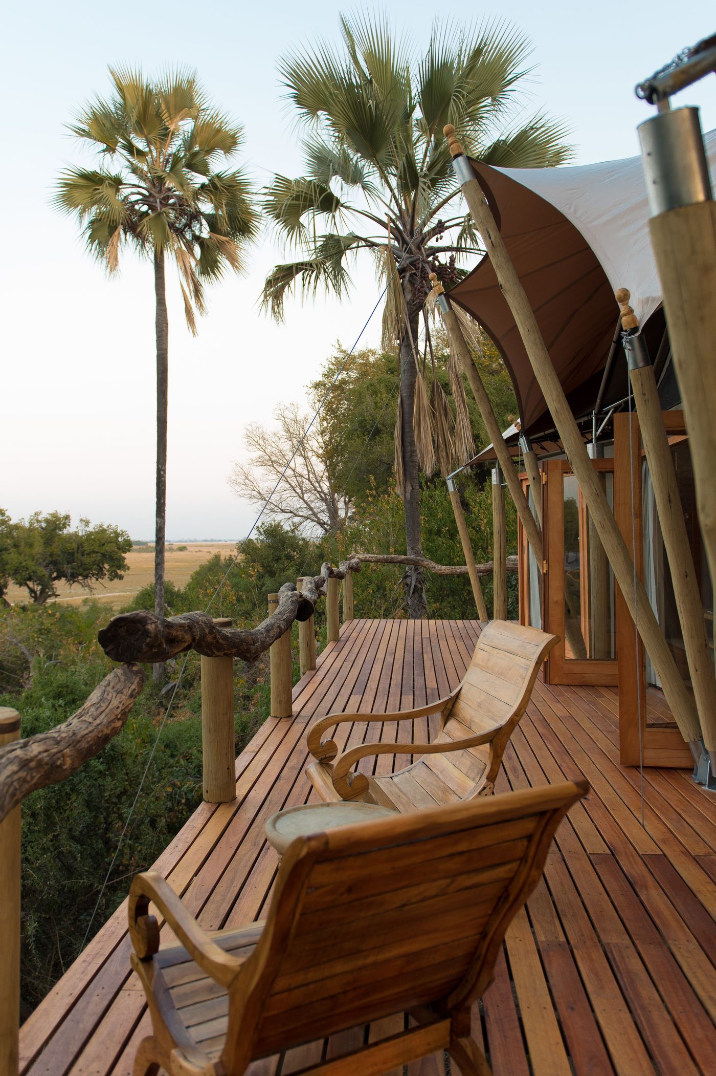 Wooden armchairs on a raised timber deck of a safari lodge with canvas tents and tall palm trees