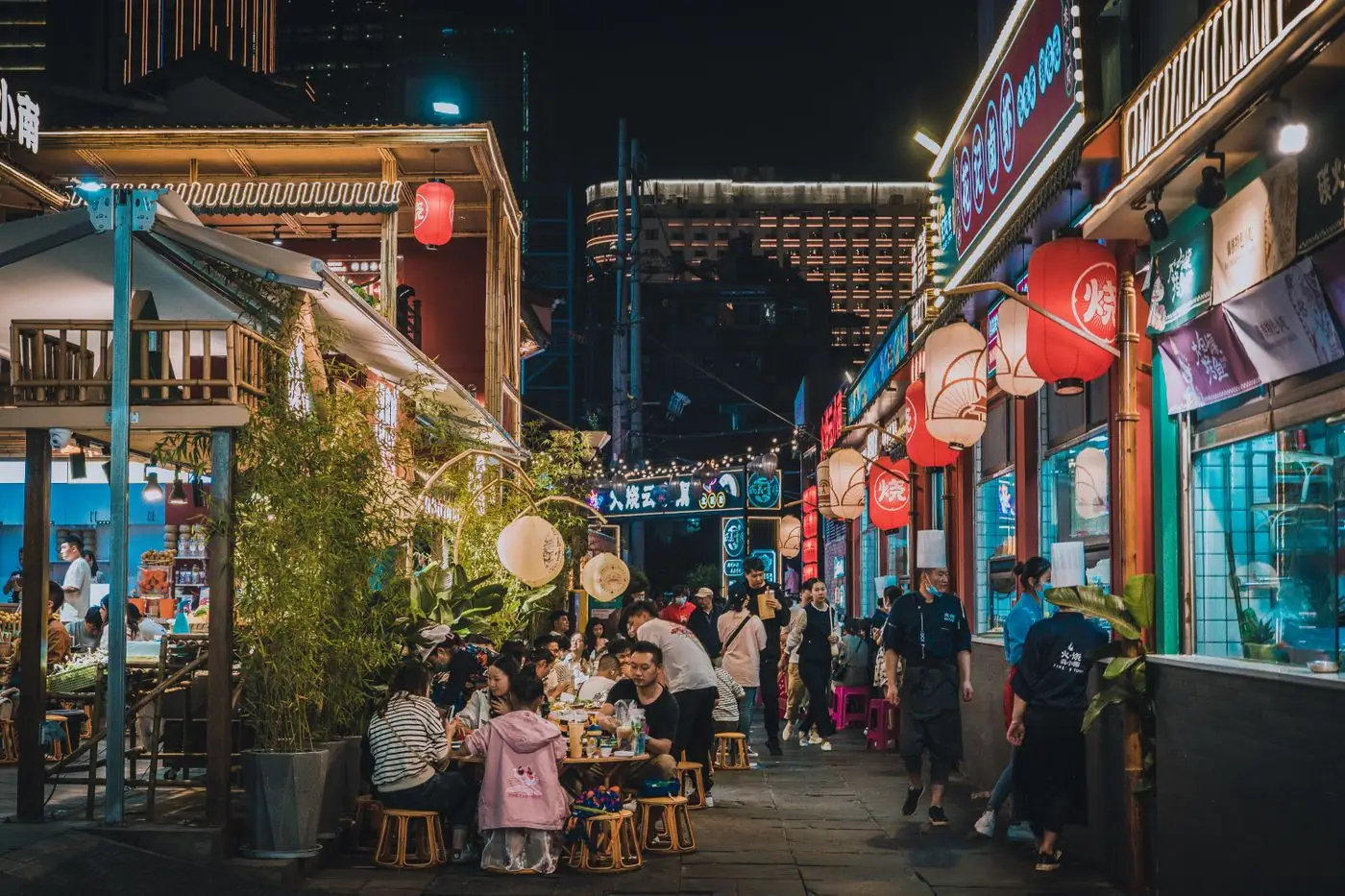 Diners at low tables on a narrow food street at night, lit by paper lanterns