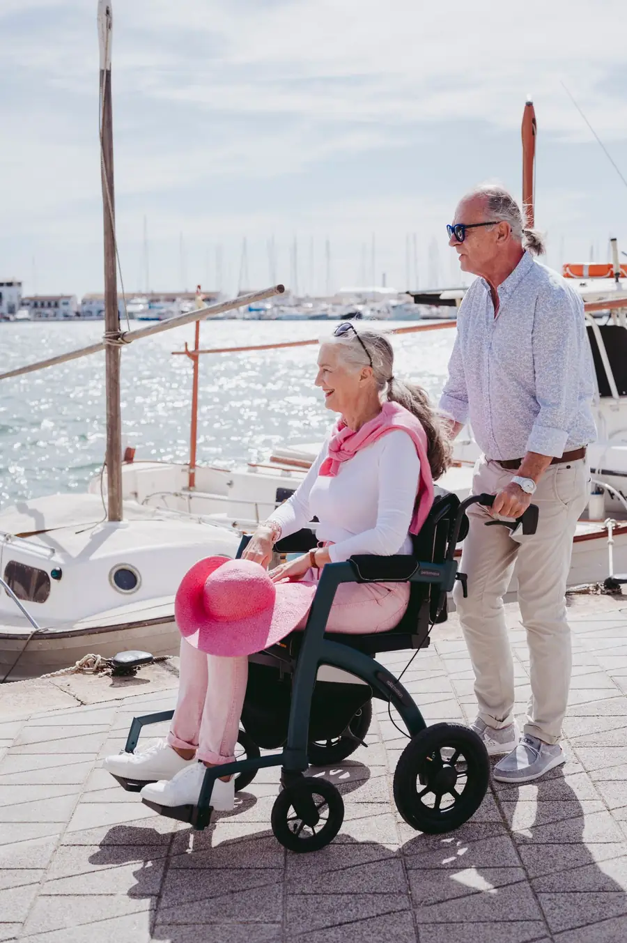 A smiling woman in a wheelchair holding a pink hat, with a man pushing her chair on a harbour promenade
